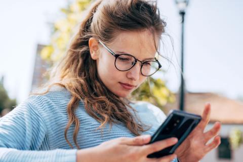 A gorgeous female in blue sweater, eyeglasses sitting on the bench reading news on smart phone outdoors on a sunny day. Happy woman takes a rest during online conversation on a mobile phone outside.