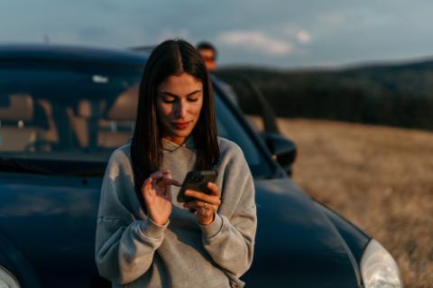 Caucasian woman leaning against her car in a grassy field, soaking in the sunset and using a phone. A man in the background.
