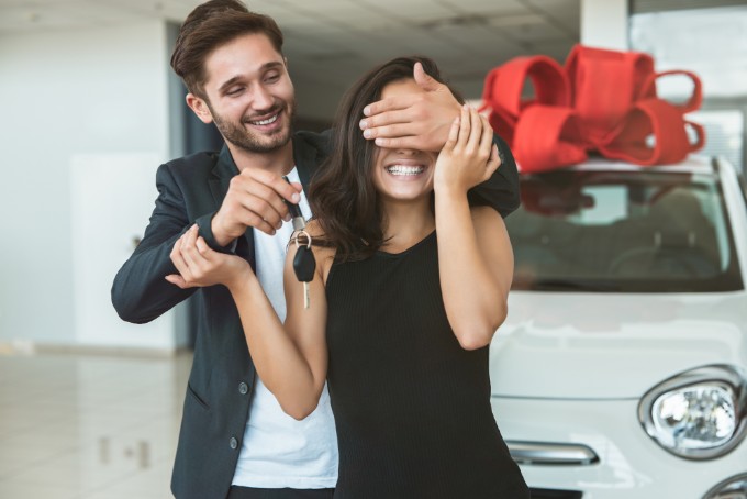 young handsome man makes surprise for his beautiful wife closes her eyes with his hands standing in dealership center buying new car for present