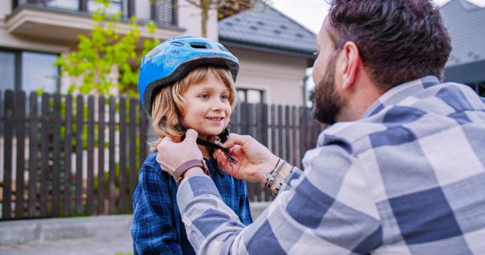 View over shoulder of father wearing helmet on head of cute little son at street. Dad teaching kid to ride a bike. Outside. Fatherhood concept.