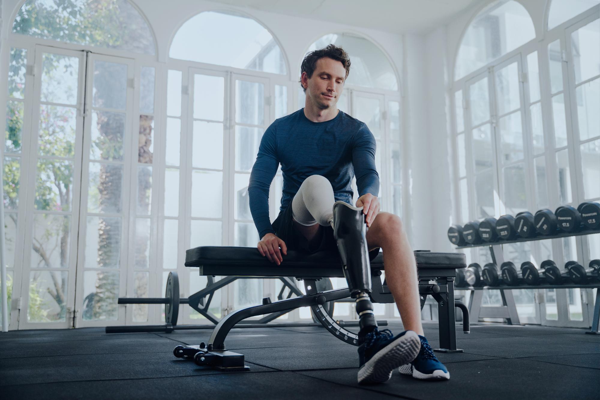 Young caucasian man in sports clothing sitting on bench press while attaching prosthetic leg at the gym