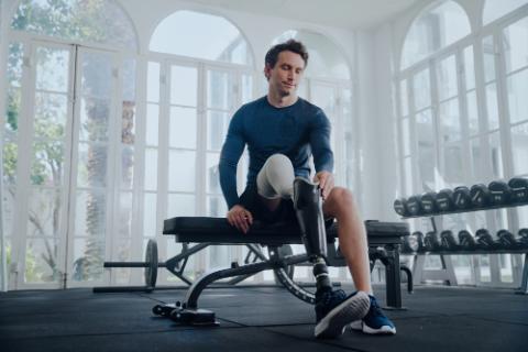 Young caucasian man in sports clothing sitting on bench press while attaching prosthetic leg at the gym